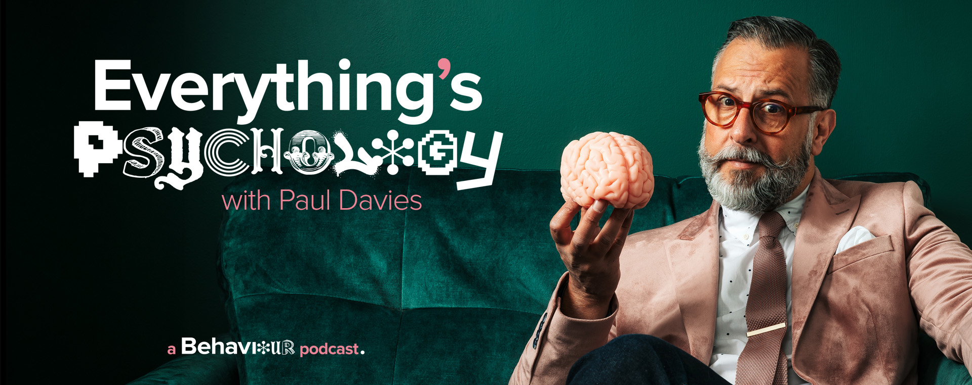 Paul Davies sat on a dark green, velvet sofa holding up a pink brain.